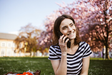 Young Student Sitting on Spring Meadow in City Park, Laughing and Talking on Smartphone - 718825534