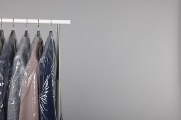 Dry-cleaning service. Many different clothes in plastic bags hanging on rack against grey background, space for text