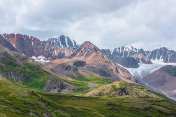 Fototapeta premium Vivid dramatic alpine scenery with green hills and rocks with view to rocky pointy peak, sharp rockies of red color, large snow mountain top, snowy range and big glacier tongue under gray cloudy sky.