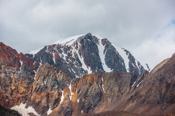 Colorful mountain landscape with beautiful large snow-capped rocky pinnacle among vivid red sheer crags in sunlight under gray cloudy sky. Dramatic  view to snow cap on multicolor mountain close up.