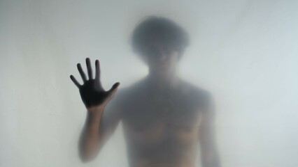 Blurred silhouette of a man with a naked torso behind a frosted curtain or glass. The man touches...