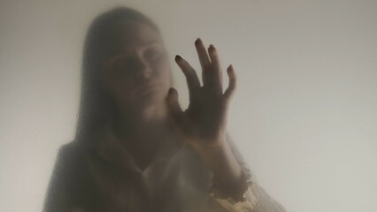 Silhouette of ghost woman in fog behind frosted curtain or glass close up. A woman is touching the...