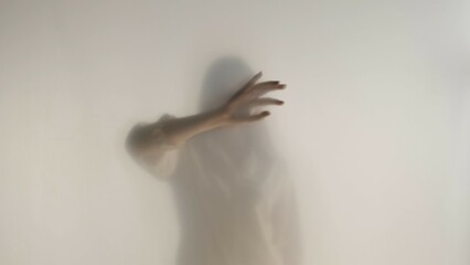 Melancholic female silhouette behind a transparent frosted curtain or glass close up. A woman is guiding her hand on the surface of the curtain.