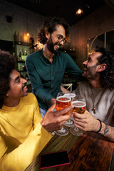 Vertical photo of multiethnic group of three male friends gathered in a bar toasting with beers, celebrating together and laughing.