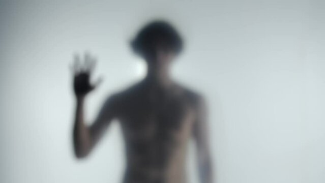 A blurred silhouette of a man with a bare torso behind a frosted curtain or glass. A man graces an invisible barrier and humbly bows his head. Ghost concept.