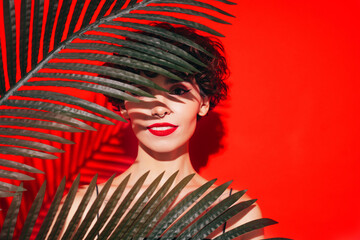 Fashion beauty portrait of young smiling brunette woman with evening stylish makeup and perfect clean skin. Sexy model with curly hair posing in studio. With red bright lips. Tropical palm leaf