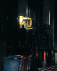 Silhouette of a person smoking under a dim light, at night, in a dark alleyway. In the blurred foreground, several large garbage cans. The lack of detail accentuates the eerie quality of the scene.