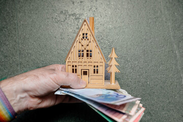 A man holds a small, wooden dream house against a stone background, accompanied by multiple euro banknotes, symbolizing aspirations for property ownership, real estate investment, or renting.