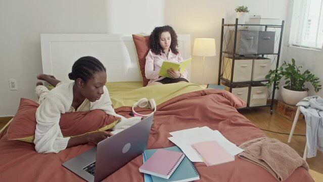 Handheld shot of female biracial roommates preparing for exam while lying in bed at home
