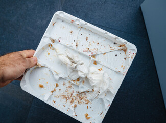 A male hand holds an empty plastic cake tray above a blue background, showcasing small traces and crumbs, remnants of an afterparty