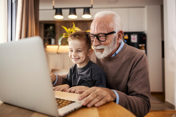 A senior casual businessman is working on a laptop from home while his grandson is helping him.