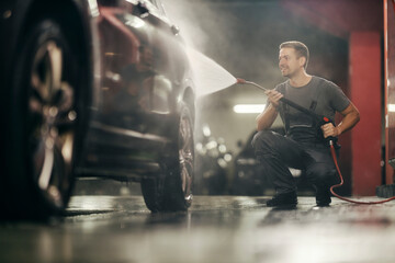 A car wash worker is crouching and washing a car with a high pressure water jet gun.