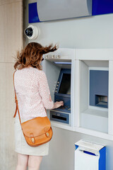 A woman is withdrawing money from an outdoor ATM as a strong gust of wind sweeps her hair upward - concept for high bank fees and withrawal of funds