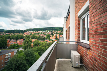 Fototapeta na wymiar Balcony of an apartment with a scenic view of a residential area. Houses with red roofs, green trees, and distant hills under a cloudy sky. There is a small AC exterior unit on the balcony.