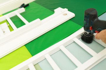 A male hand with a drill assembles a child bed, focusing on attaching the protective grill to prevent the baby from falling off the bed