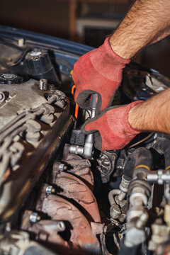 The mechanic is screwing back the exhaust manifold into the car using a ratchet wrench. Open hood, valve cover, spark plug wires, and dipstick. Red protective gloves.