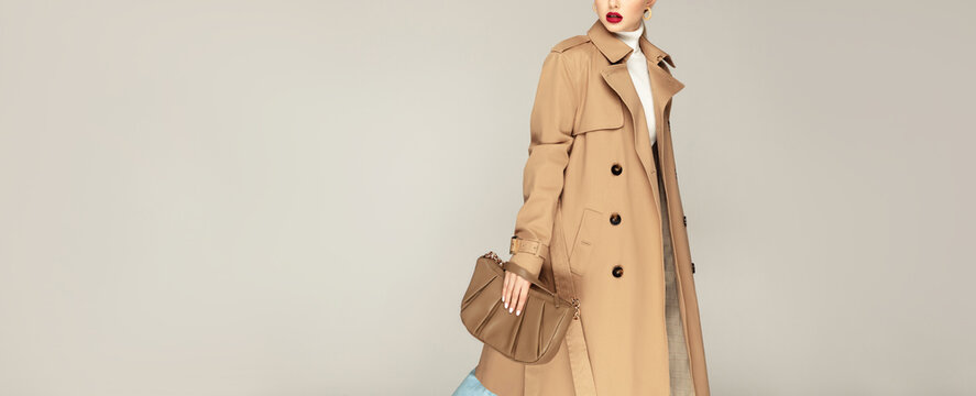 Autumn clothing shopping concept. Fashionable young model posing against grey background dressed in trench coat and holding stylish  bag.