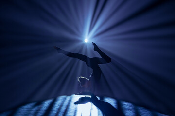 Female gymnast on dark background of studio with backlight. Acrobatic girl performing handstand. Modern choreography and acrobatics creative advertising concept.