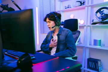 Young Asian man professional gamer wearing a jeans jacket sits on a chair and grabs his chest...