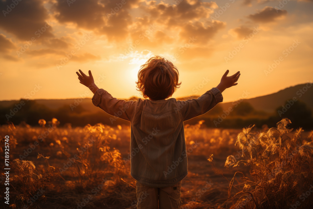 Wall mural A little boy raises his hands above the sunset sky, enjoying life and nature. Happy kid on a summer field looking at the sun. Silhouette of a male child in the sun. Fresh air, environment concept.