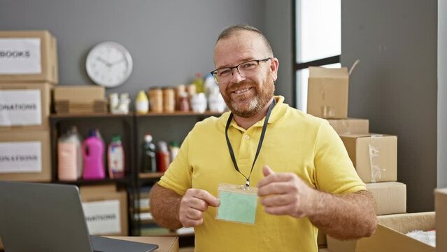 Handsome adult caucasian man, a confident charity volunteer, smiling and pointing at his identification id while working in a community center, catches you on camera indoors.