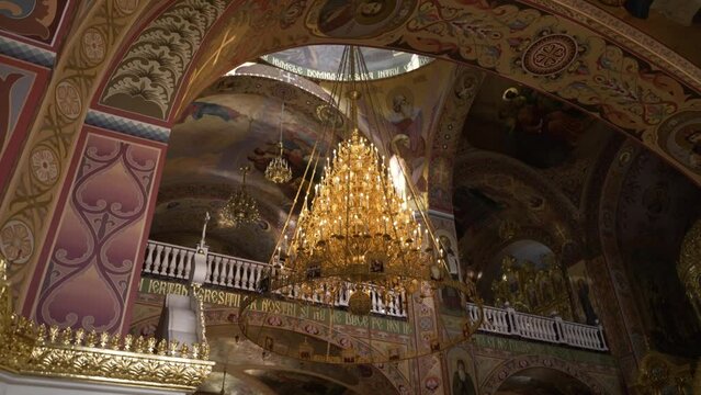 A magnificent chandelier descends from the vaulted ceiling of an Orthodox church, set against a backdrop of detailed frescoes and ornate patterns.