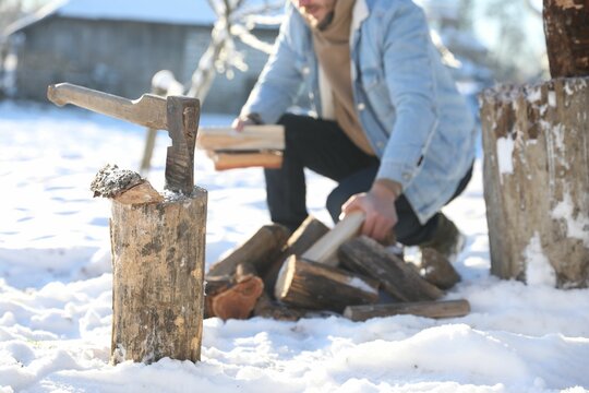 Man taking logs from pile outdoors on winter day. Metal axe in wooden log on snow, selective focus