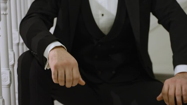 a man in a formal black suit is seated, attentively fastening his cufflinks. His sleeves are crisp white, contrasting with the suit, signifying formality and preparation for a significant event.