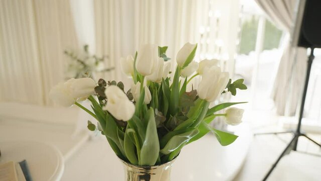 A bouquet of white tulips with lush green leaves and eucalyptus branches in a glass vase, set against a softly lit room with white drapes.