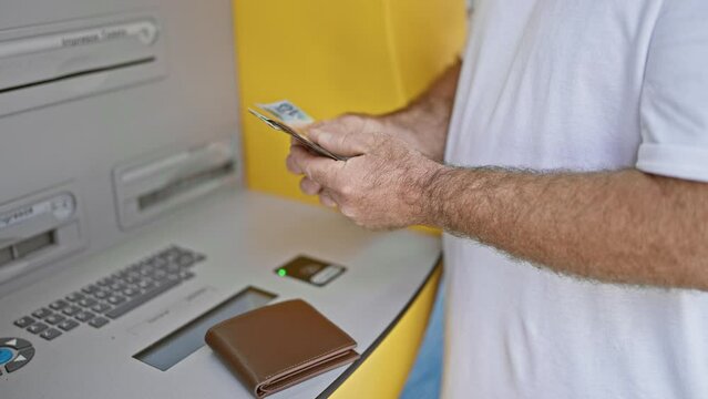 Handsome middle-aged caucasian man holding australian dollars, caught mid-transaction at urban street atm. outdoor banking technology in action, person withdrawing cash in australia.