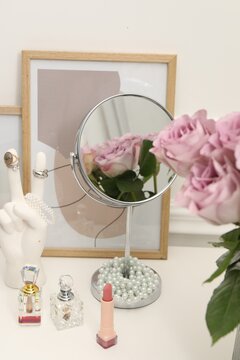 Mirror, cosmetic products, perfumes and vase with pink roses on white dressing table