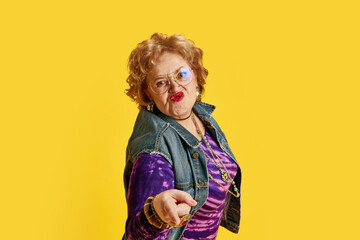 Senior woman puckering lips in round glasses dressed denim jacket, purple T-shirt with gold jewelry against yellow background. Concept of empowerment of older adults, active seniors in modern life.