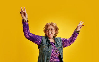 Elderly woman making peace sign and laughing dressed denim jacket and vibrant purple top. Happy...