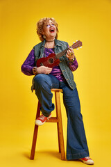 Joyous senior woman energetic playing ukulele seat on stool dressed casual denim and red shoes....