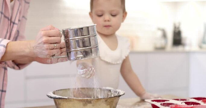 happy mother and little child girl sieving flour into bowl. baby girl is playing with flour in the kitchen.