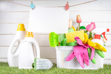Spring cleaning background. Set of cleaning supplies, bottles, accessories and stuff with spring flowers in basket on white wooden green garden grass background copy space