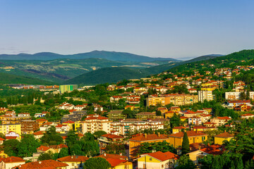 Fototapeta na wymiar panoramic landscape view of green summer city with yellow houses, green forest and mountains above and amazing hills and cloudy sky on background