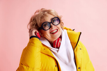 Cheerful senior woman enjoying listening music in headphones dressed yellow puffer jacket, white...