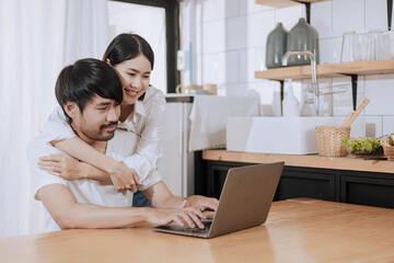 Happy love Asian young man and woman using laptop together in the kitchen relaxing at home.