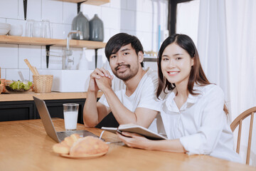 Happy love Asian young man and woman using laptop together in the kitchen relaxing at home.