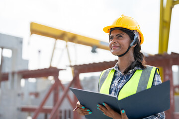 Asian woman engineer holding document smiling at construction site. Confident female Indian wearing protective helmet and vest working in factory making precast concrete wall for real estate housing.