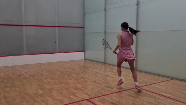 Squash player hits ball against white wall marked with red lines. Young woman playing with a racket on court