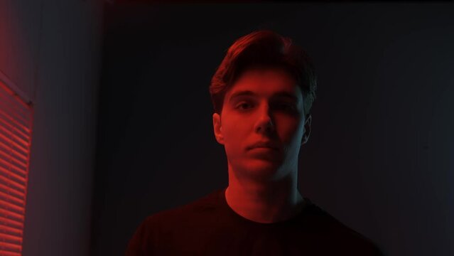 Portrait of male model in dark studio. Young man in t-shirt standing near window with red light behind jalousie looks away and turns at camera.