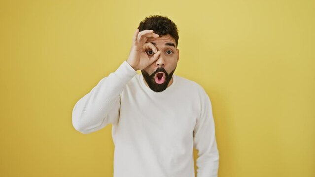 Shocked young man stands in disbelief, eyes peering through ok sign on fingers. his surprise etched across his face, a sight against yellow isolated backdrop. quirky, confident yet taken aback.
