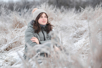 Winter portrait of young woman baby face in winter clothes sitting on field on frosty day. Pretty girl on nature