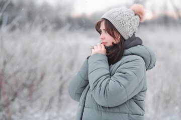 young woman in hat warms her hands with breath, cute girl walking on in frosty day, winter walk