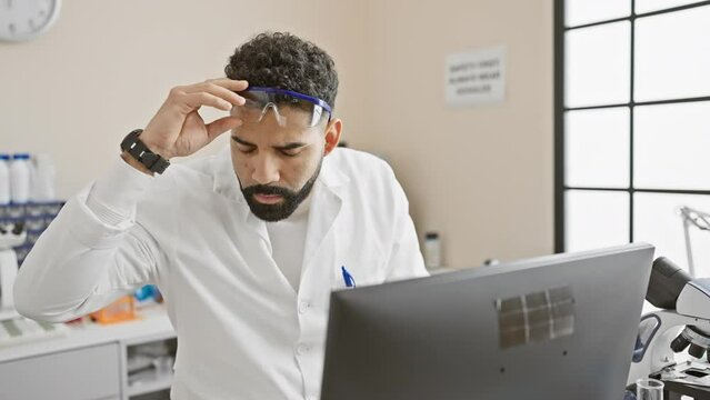 A focused hispanic man with a beard wearing safety goggles and a lab coat analyzes data on a computer in a laboratory setting.