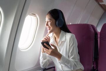 Cheerful Asian woman passenger in headphone watching online movie during intercontinental flight in cabin of aircraft, happy traveler using wifi connection on board - obrazy, fototapety, plakaty