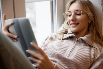 Young woman with braces wearing beige sweater with glasses, smiling with reading tablet by window. Smiling patient. Concept of beauty and medicine, dental care, malocclusion, orthodontic health. - obrazy, fototapety, plakaty
