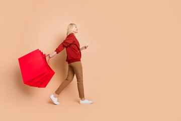 Full length photo of cute good mood lady dressed red pullover walking holding bargains ordering device empty space isolated beige color background